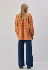 Chemise surdimensionnée à carreaux jaunes avec des accents roses, manches longues et ourlet arrondi, associée à un pantalon en denim bleu à jambes larges.