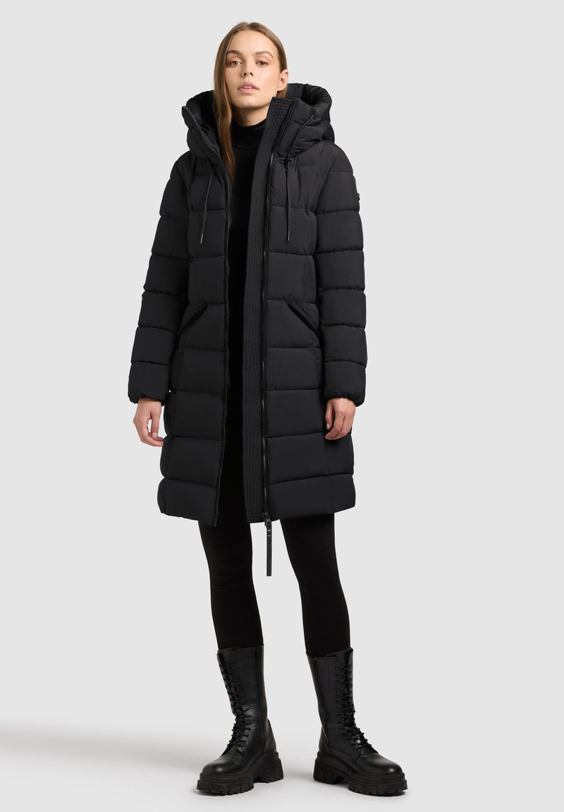 Piumino nero con cappuccio, caratterizzato da sezioni trapuntate orizzontali, una chiusura frontale a zip e due tasche laterali. Indossato con leggings neri e stivali militari.