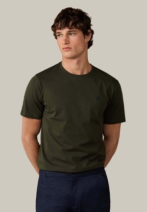 Joven con pelo corto y rizado, vestido con una camiseta verde oscuro y pantalones azul marino, de pie con las manos detrás de la espalda, mirando hacia un lado.