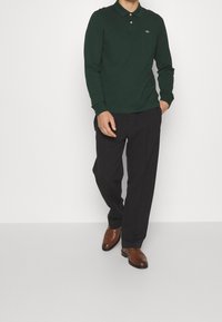 Dark green long-sleeve polo shirt with a collar and buttons, paired with black trousers and brown dress shoes. Minimal logo detail visible.