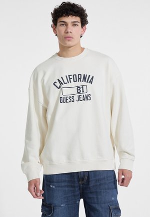 Svetlo kremasti dukserica s opuštenim krojem, s tamnoplavim natpisom "CALIFORNIA 81 GUESS JEANS" u središtu. Rebrasti rukavi i donji rub.