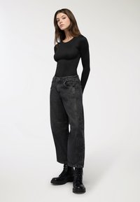 Czarna bluzka z długim rękawem zestawiona z ciemnoszarymi, luźnymi dżinsami o wysokim stanem. Modelka nosi czarne, sznurowane buty wojskowe. Prosty, nowoczesny design.