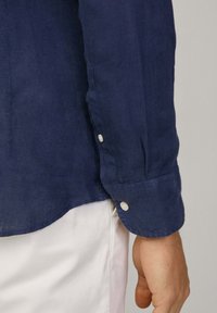 Camicia in lino blu navy a maniche lunghe con polsini a bottoni, caratterizzata da una sottile trama e un orlo curvo. Ingrandimento della manica e dell'area del polso.