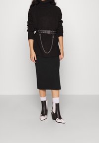 Svart stickad polotröja med en nitarbälte, svart blyertskjol och svarta ankelskor med vita accenter och mönstrade detaljer.