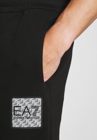 Svarta joggers med en hand i sidfickan, med en logotyp patch på en grå och svart mönstrad bakgrund och vit text.