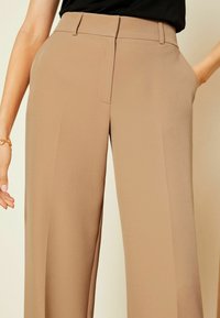 Pantaloni beige a gamba larga realizzati in tessuto morbido, con frontale piatto, passanti per cintura e tasche laterali. Nessun motivo o dettaglio visibile.
