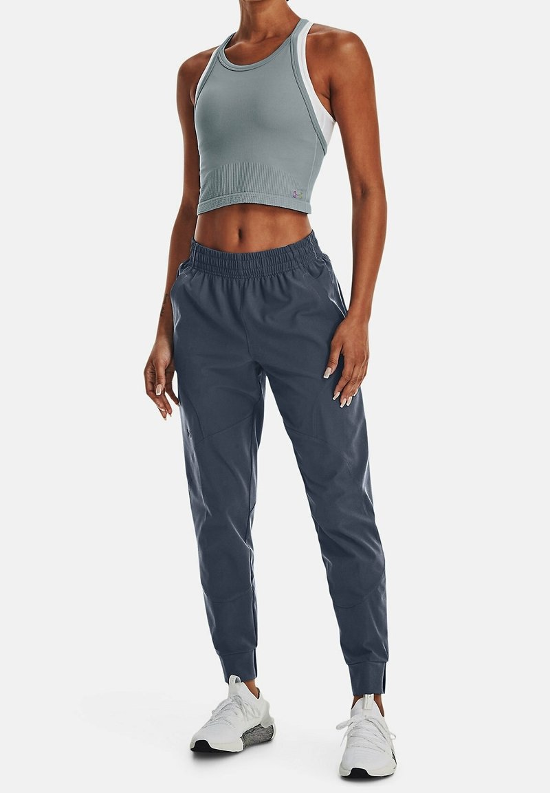 Woman wearing gray cropped tank top, dark blue jogger pants, and white athletic shoes, standing with hands relaxed by sides.