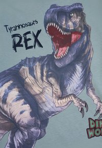 T-Shirt mit einem großen, detaillierten Grafik eines Tyrannosaurus Rex mit offenem Maul auf einem hellblauen Hintergrund. Der Text lautet "Tyrannosaurus REX."