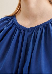 Blusa blu con scollatura rotonda e tessuto arricciato, con pieghe verticali che aggiungono texture. Materiale liscio con una leggera lucentezza.