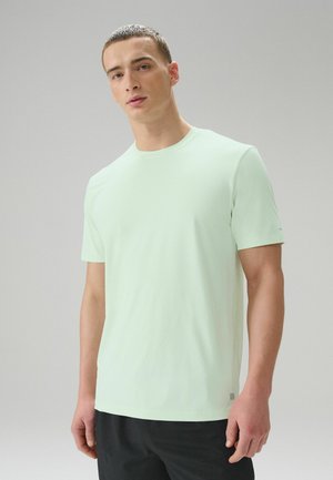 Young man with short hair wearing a light mint green short-sleeve shirt and black pants against a plain gray background.