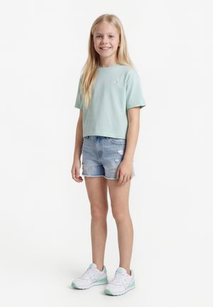 Fille blonde souriante portant un t-shirt vert menthe, un short en jean clair et des baskets blanches avec des accents verts menthe, debout devant un fond blanc uni.