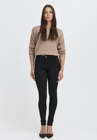 Bežinis megztinis su laisvu siluetu, derinamas su juodomis liesomis džinsomis. Rudi bateliai su smailiu pirštu užbaigia aprangą. Neutrali foninė aplinka.