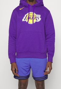 Fioletowa bluza z kapturem z logo Los Angeles Lakers, wyposażona w przednią kieszeń i sznurki do ściągania. W zestawie niebieskie, perforowane spodenki z czarnymi paskami.