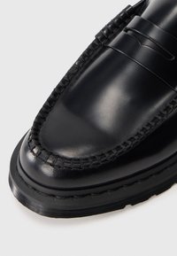 Close-up de sapato loafer de couro preto polido com solado grosso costurado e detalhes à mão em estilo mocassim na área do dedão.