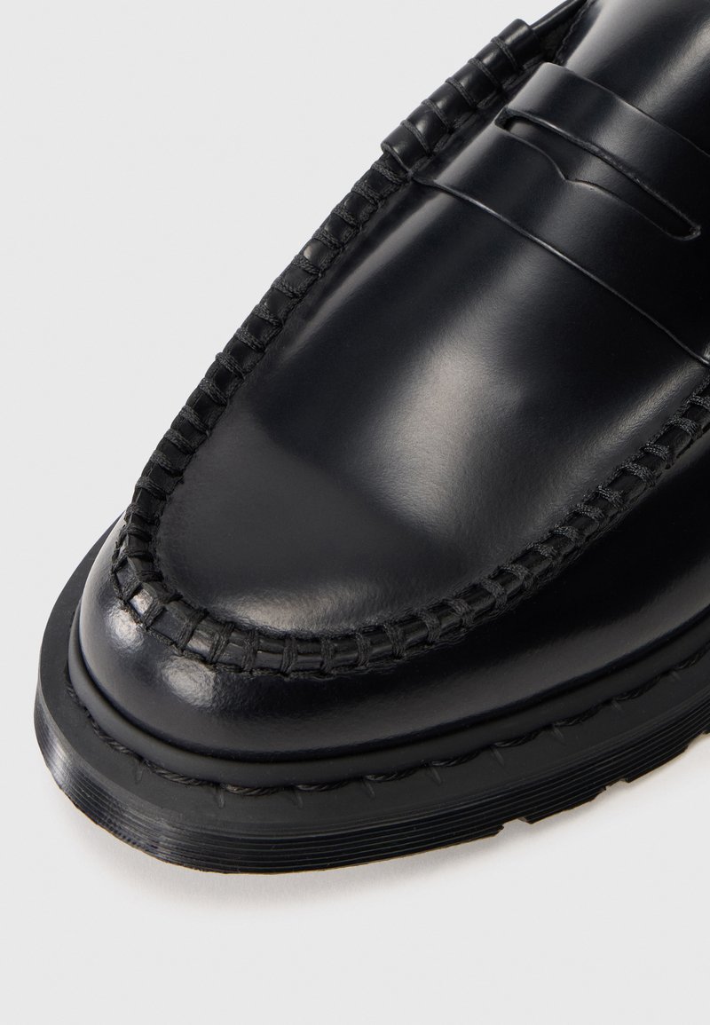 Primo piano di una scarpa loafer in pelle nera lucida con suola spessa cucita e dettagli cuciti a mano in stile mocassino nell'area delle dita.