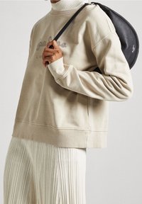 Sudadera beige con logo bordado, cuello de canalé y mangas largas, combinada con pantalones blancos con pliegues y un pequeño bolso negro redondeado.