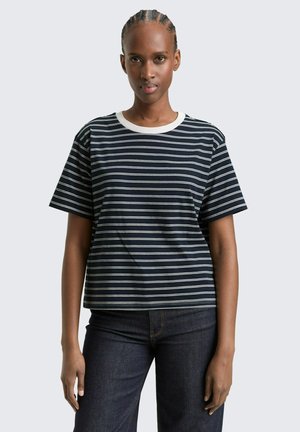 Frau mit geflochtenem Haar, die ein schwarz-weiß gestreiftes T-Shirt mit horizontalen Streifen und eine dunkle Jeans trägt, steht vor einem schlichten hellgrauen Hintergrund.