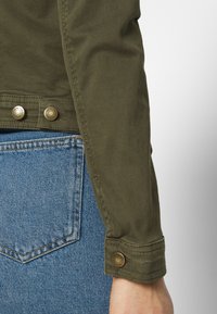 Olivgrüne Jacke mit hochgekrempelten Ärmeln, versehen mit Messingknöpfen und einem klaren, maßgeschneiderten Design. Kombiniert mit hellblauen Jeans.