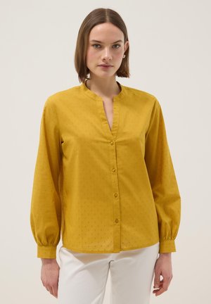 Femme aux cheveux bruns courts portant un chemisier à manches longues moutarde avec des boutons et un pantalon blanc, devant un fond clair uni.