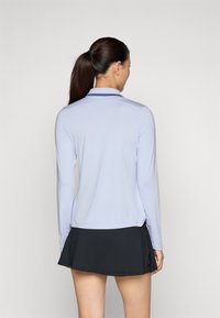 Long-sleeve light blue polo shirt with a collar and side slits, paired with a black skirt. Smooth fabric, fitted design, minimal detailing.