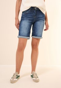 Shorts en denim bleu moyen avec des revers roulés, dot�és d'une fermeture à bouton à l'avant et d'un design à cinq poches. Assortis à des baskets avec des accents verts.