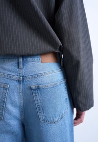 Άτομο που φοράει ανοιχτό μπλε τζιν με πίσω τσέπες και σκούρο γκρι σακάκι με ρίγες, το χέρι αναπαύεται στο πλάι.