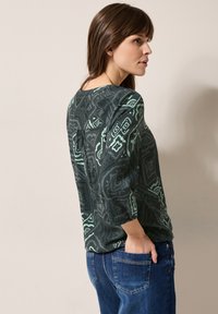 Haut à manches longues vert foncé à motifs avec des dessins géométriques vert clair, col rond et coupe décontractée avec des manches froncées.