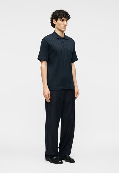 Νεαρός άνδρας με κατσαρά μαλλιά, φορώντας σκούρο μπλε ριμπ κοντομάνικο πόλο και αντίστοιχο φαρδύ ριγέ παντελόνι με μαύρα παπούτσια.