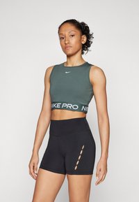 Zeleni Nike skrajšan top z belim logotipom in črnim Nike Pro pasom, v kombinaciji z visokimi črnimi kratkimi hlačami z izrezanimi detajli.