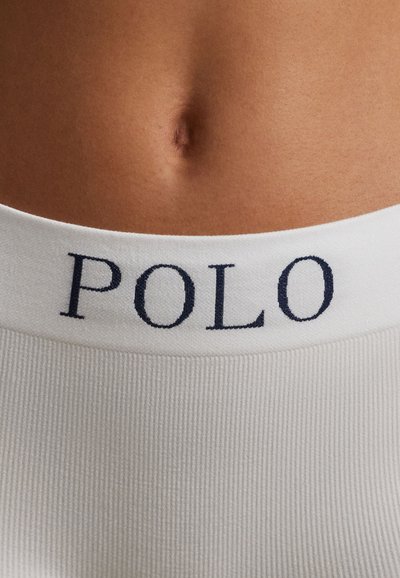 Zbliżenie na środkową część ciała osoby noszącej białą odzież z prążkowanej tkaniny, z paskiem w talii oznaczonym ciemnymi literami "POLO".