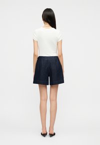 Mujer de espaldas, vestida con una camiseta blanca de manga corta, pantalones cortos de mezclilla oscura, zapatos negros de tacón y un collar de perlas.