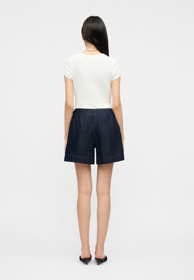 Γυναίκα που στέκεται γυρισμένη πλάτη, φορώντας λευκό τοπ με κοντά μανίκια, σκούρα τζιν σορτς, μαύρα παπούτσια με τακούνι και κολιέ με πέρλες.