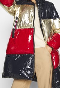 Vadderad jacka med färgblock: glansigt guld, röd och marinblå sektioner. Har en zip-ficka på sidan och en hög krage. Slät, glänsande textur.