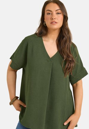 Kvinde med langt brunt hår iført en løs, kortærmet, olivengrøn V-hals top og blå jeans, med den ene hånd i lommen og armbånd om håndleddet.