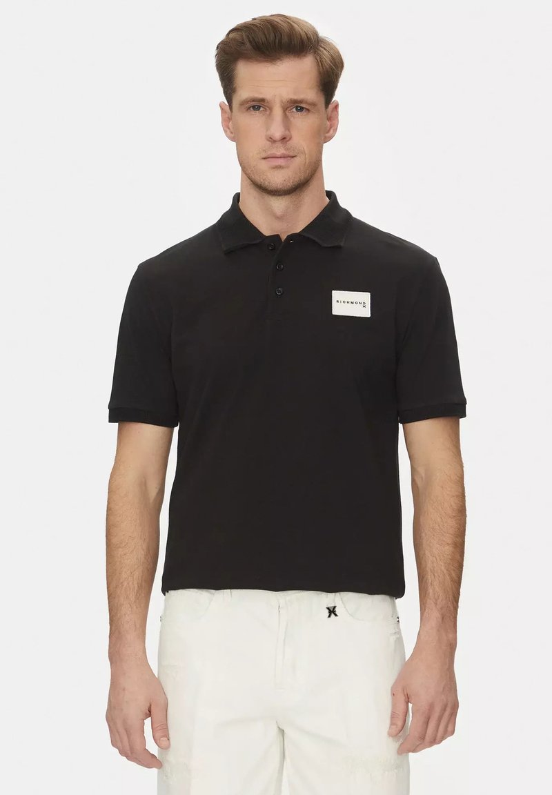 Polo shirt nera in cotone con colletto, maniche corte, patta con tre bottoni e una toppa rettangolare bianca sul petto.