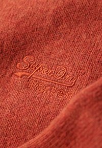 Tessuto rosso texture con logo "SuperDry" ricamato in una tonalità più chiara. La vista ravvicinata mostra i dettagli della tessitura e la morbidezza del materiale.