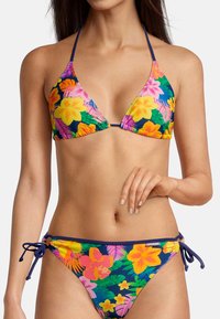 Haut de bikini triangulaire et bas de bikini noué dans un motif floral vibrant avec du rose, du jaune et du vert sur un fond bleu marine. Texture de tissu lisse.
