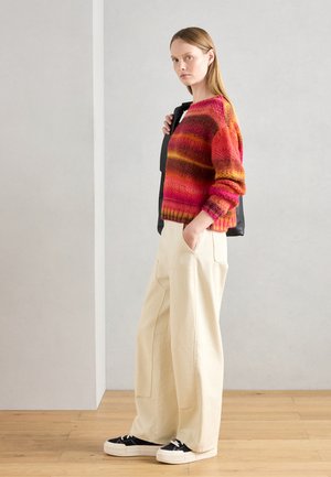 Pull en tricot avec des rayures rouges, roses et oranges, associé à un pantalon large crème et des baskets noires avec des semelles blanches.