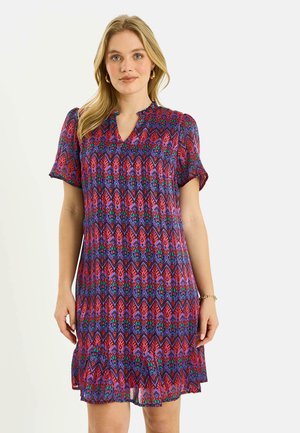 Mujer con vestido de manga corta hasta la rodilla con un patrón geométrico colorido en rojo, púrpura y azul, de pie frente a un fondo liso.