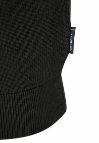 Schwarzer Strickpulloverstoff mit gerippter Textur. Hat ein dezentes Logo-Etikett in Blau mit weißen Text, das an der Seitennaht platziert ist.