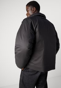 Giubbotto a sacco nero con colletto alto, caratterizzato da una texture liscia e una vestibilità oversize. Il modello indossa pantaloni scuri, presentando uno stile rilassato.