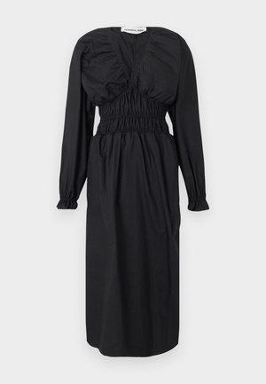 Kvinne står med armene i kors iført en svart midi-kjole med lange ermer og svarte hælede mules mot en ensfarget grå bakgrunn.