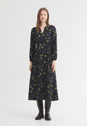 Frau trägt ein langärmeliges, marineblaues Kleid mit grünem Blumenmuster und schwarzen kniehohen Stiefeln und steht vor einem weißen Hintergrund.