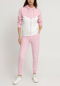Haut à capuche zippé rose et blanc avec un design en chevron, associé à un jogger rose assorti. Fabriqué en tissu doux, il présente des poignets côtelés et des poches latérales.