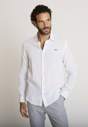 Homme aux cheveux bouclés portant une chemise blanche boutonnée et un pantalon gris clair, debout avec une main dans la poche, regardant vers la droite.