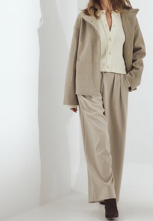 Mujer con chaqueta de lana beige, cárdigan de punto color crema con botones, pantalones beige holgados y botas oscuras caminando sobre fondo blanco.