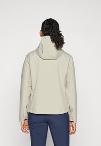 Donna con capelli ricci raccolti in una coda di cavallo, indossa una leggera giacca con cappuccio beige chiaro e pantaloni blu navy, rivolta di spalle su uno sfondo neutro.