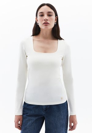 Haut à manches longues blanc avec un décolleté carré, fabriqué dans un tissu lisse. Présente un détail de logo subtil à la taille. Associé à un jean bleu.