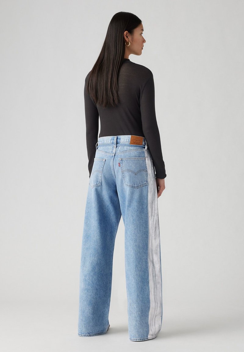 Jeans a gamba larga blu chiaro con un pannello laterale sbiadito. Presentano una toppa in pelle sul retro e un design classico a cinque tasche. Indossati con una maglietta nera attillata.