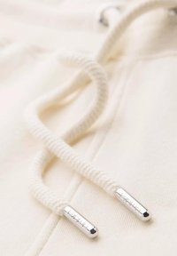 Cordon de sudadera en algodón crema, con textura retorcida, con terminales de metal plateado etiquetados "marca". Enfoque en primer plano del material y los detalles.